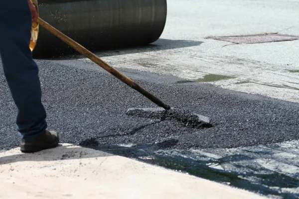 This image shows a worker using a rake to spread asphalt. The worker is wearing dark pants and work boots. Freshly laid, dark gray asphalt is being smoothed out. In the background, part of a large roller machine is visible, which is likely used to compact the asphalt. The surrounding area appears to be a paved surface undergoing repair or new construction.