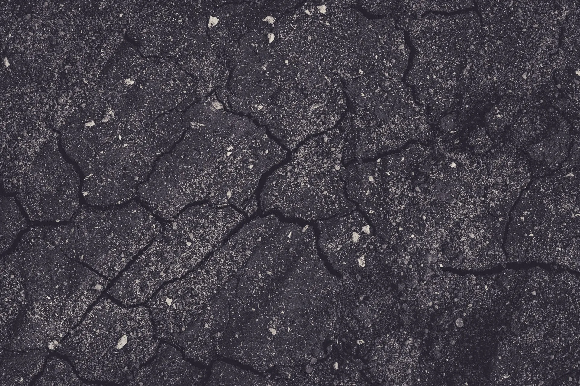 The asphalt surface is dark gray or black and is heavily fractured with a network of irregular cracks running across it. Small pieces of lighter-colored aggregate are visible embedded within the asphalt. The texture is rough and uneven due to the cracking. This suggests that the asphalt is old, weathered, and possibly in need of repair or resurfacing.