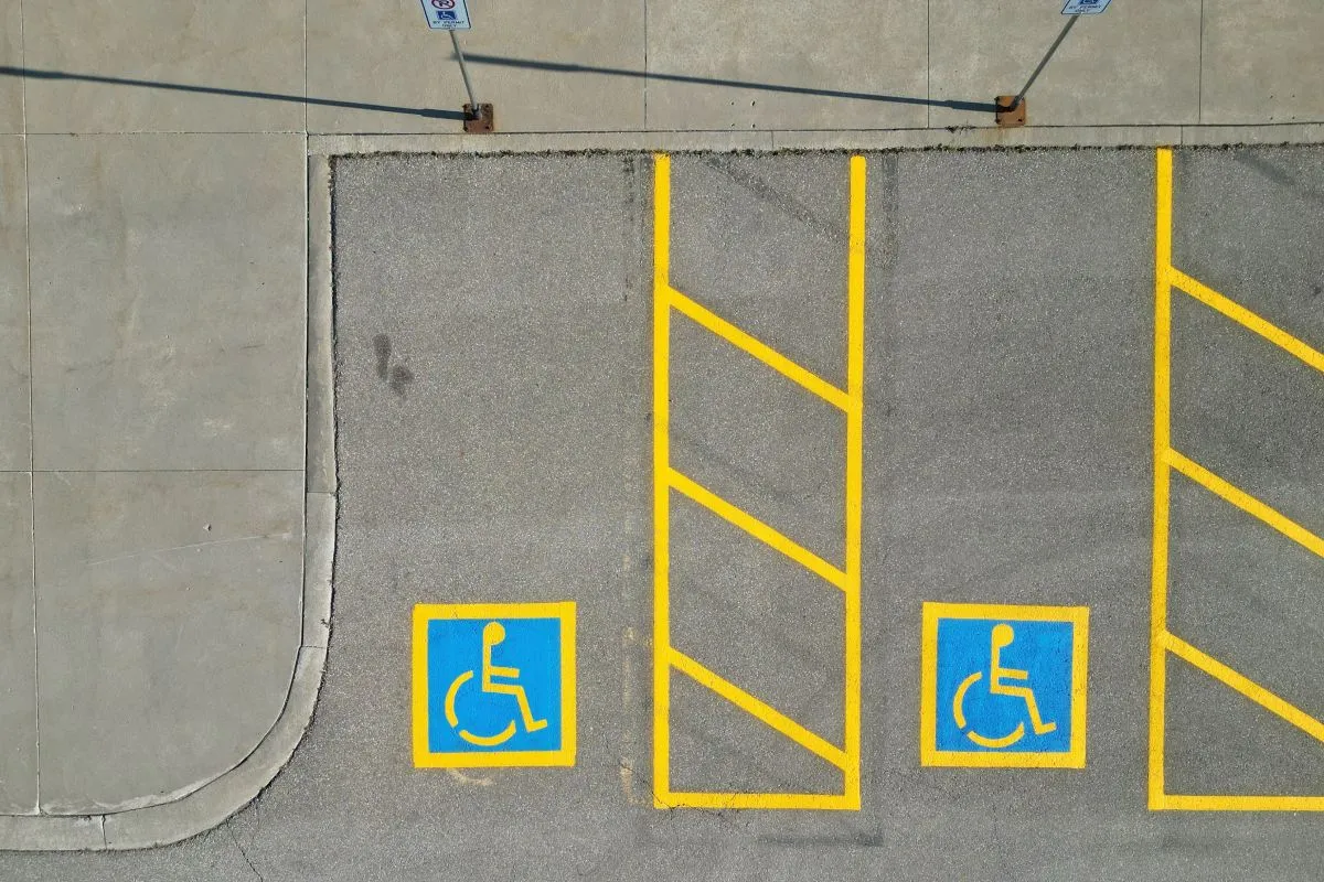 This is an aerial view of a parking lot section with designated handicapped parking spaces. The spaces are marked with blue painted squares containing the universal wheelchair symbol in yellow. Yellow lines delineate the parking stalls and adjacent access aisles with diagonal stripes. Two parking signs on poles are visible at the top of the image, further indicating handicapped parking. The parking lot surface is asphalt, and a concrete walkway runs along the top edge.