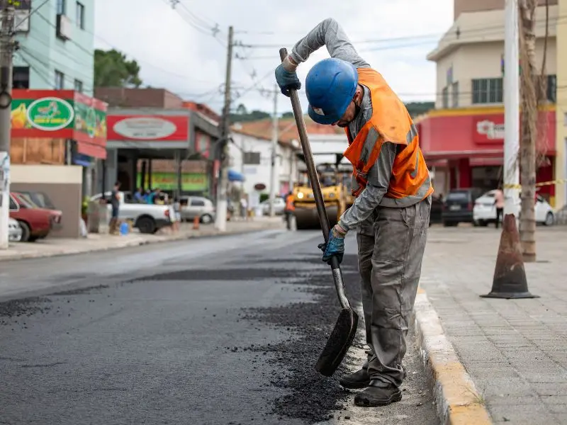 Worker: A construction worker wearing a blue hard hat, an orange safety vest, gray long-sleeved shirt, gray pants, and gloves is using a shovel to spread fresh asphalt on a section of the road.
